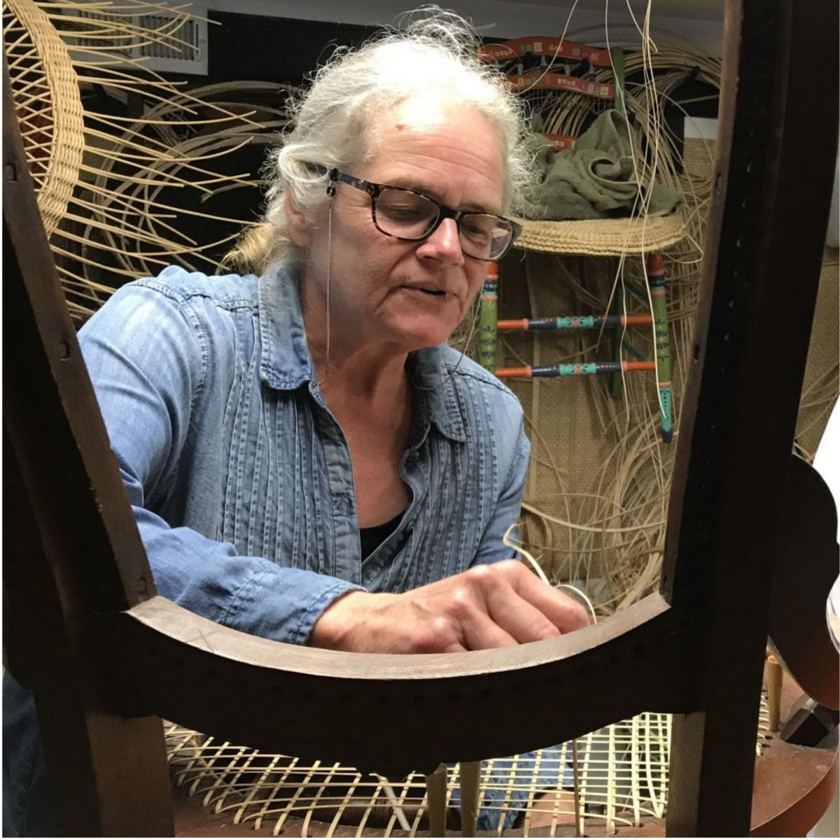 Image of a woman behind a wooden chair frame. She is wearing glasses and her hands are weaving rattan cane into the chair seat