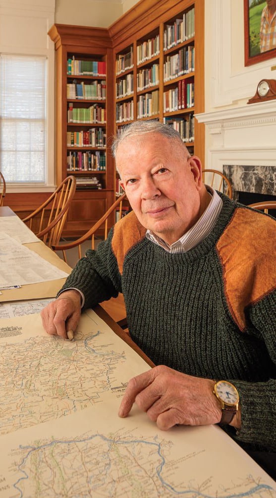 Cartographer Eugene Scheel is seated at a table, his hands are resting on some of his illustrated historical maps. Books fill the shelves behind him.